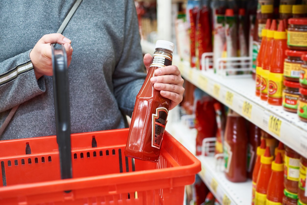 A person holding a bottle of ketchup and a red shopping basket stands in front of a grocery store shelf filled with various bottles of sauces and condiments.