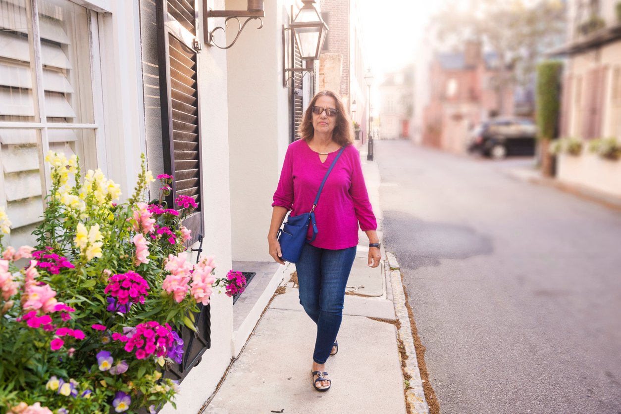 A woman wearing a bright pink top, blue jeans, and sandals walks along a quiet street lined with flowers and historic buildings on a sunny day. She has a blue crossbody bag and sunglasses.