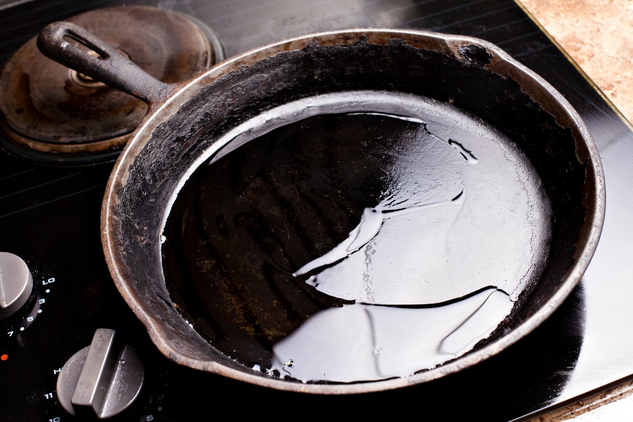 A black cast iron skillet with a shiny layer of oil on its surface sits on a stovetop with a visible burner and control knobs.