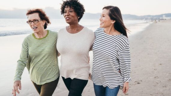 Three women walk arm-in-arm along a beach, smiling and wearing casual striped tops. The ocean and sky are visible in the background, creating a relaxed and joyful atmosphere.