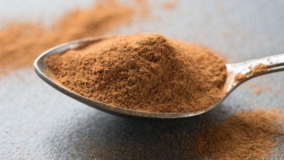 A close-up of a spoon heaped with ground cinnamon, with some cinnamon powder scattered on the dark surface beneath it.