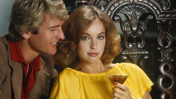 A man with blond hair in a brown jacket leans in, smiling, towards a woman with wavy brown hair in a bright yellow dress, who looks at the camera while holding a cocktail glass. Ornate wooden chair is in the background.