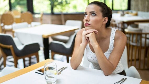 A woman sits alone at a restaurant table with her hands clasped under her chin, looking upwards. A glass of water with lemon, phone, fork, and knife are on the table. The restaurant is empty and bright.
