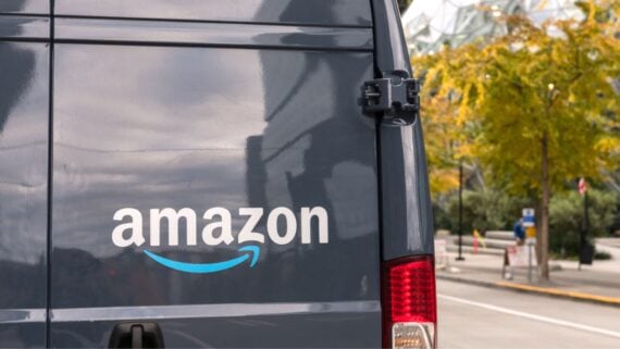 A close-up of the back of a dark blue Amazon delivery van parked on a street, with the Amazon logo clearly visible. Trees with yellow leaves and buildings are in the background.