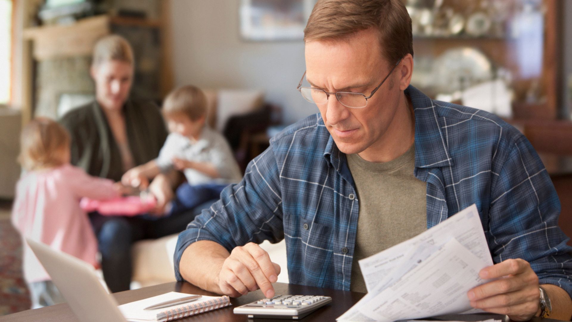 A man wearing glasses uses a calculator and checks bills at a table, while a woman and two young children play in the background.