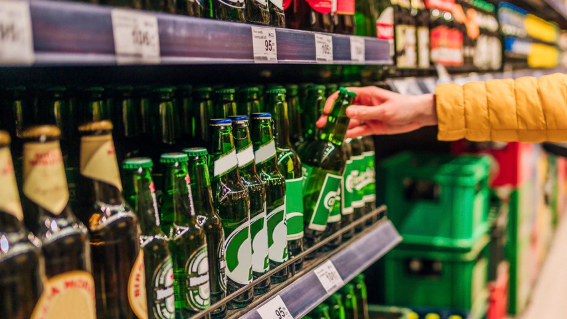 A person in a yellow jacket selects a green glass bottle of beer from a shelf in a store, surrounded by various other bottled drinks and crates.