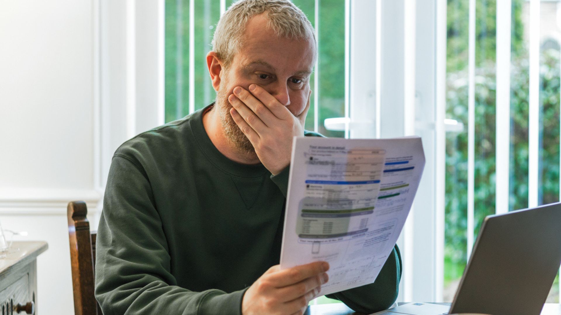A man with a concerned expression sits at a table, holding a sheet of paper with financial information, and covering his mouth with his hand. A laptop is open in front of him, and window blinds are visible in the background.