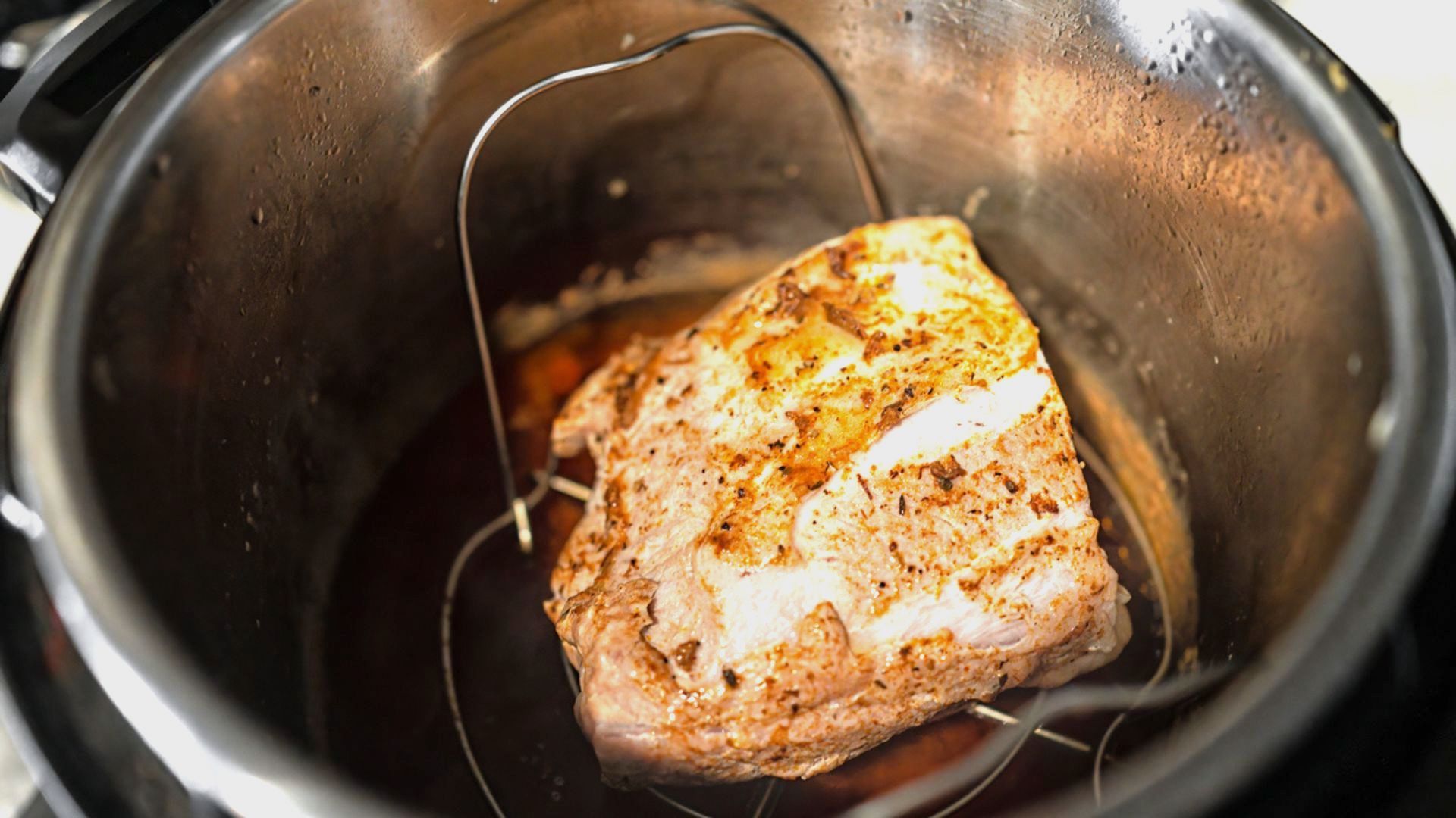 A cooked, seasoned piece of meat sits on a metal rack inside an Instant Pot with juices collected at the bottom.