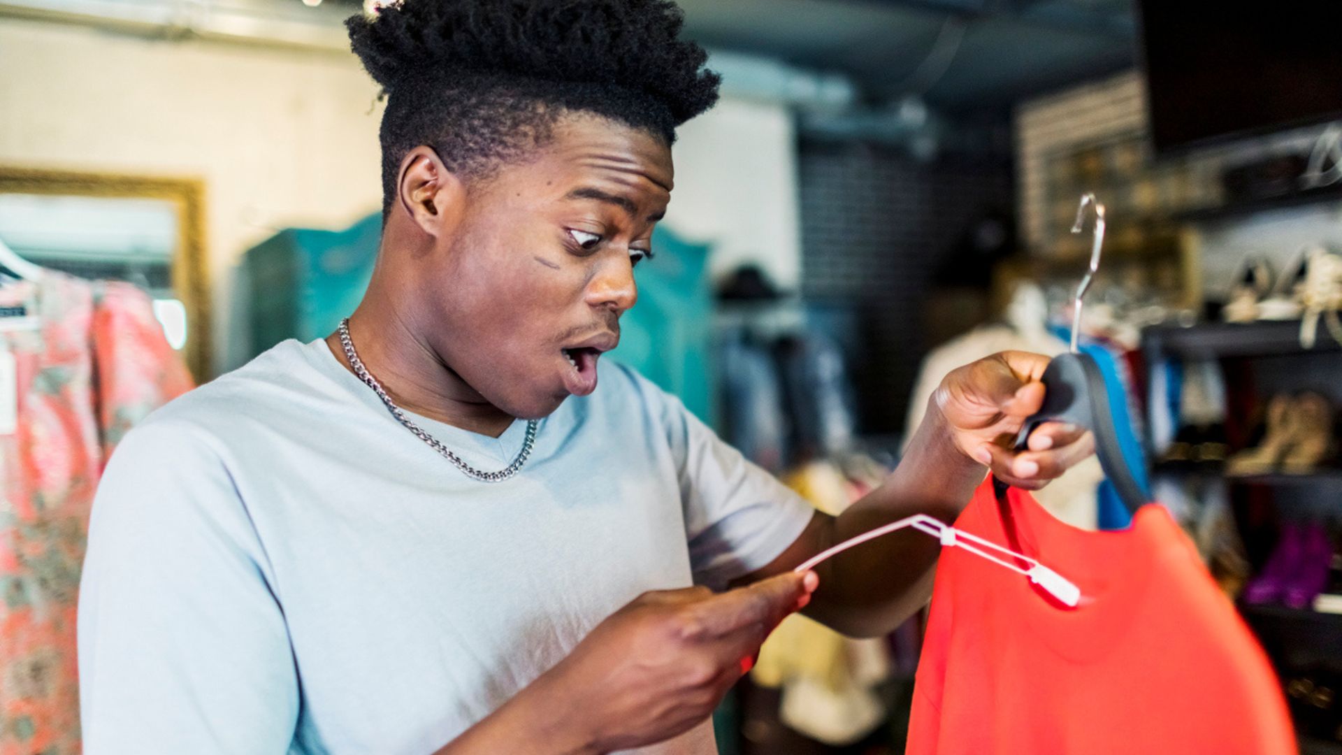 A young man with a surprised expression holds up a red dress on a hanger while looking at its price tag in a clothing store.