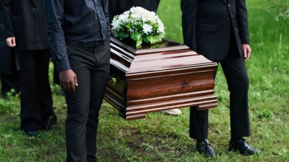 Pallbearers dressed in black carry a wooden casket adorned with a white floral arrangement across a grassy area during a funeral.