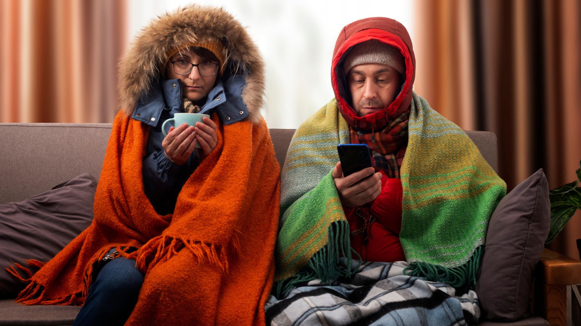 Two people sitting on a couch bundled up in winter coats, hats, and layered blankets. One holds a mug and looks ahead, the other checks a smartphone. Both appear cold, with serious expressions.