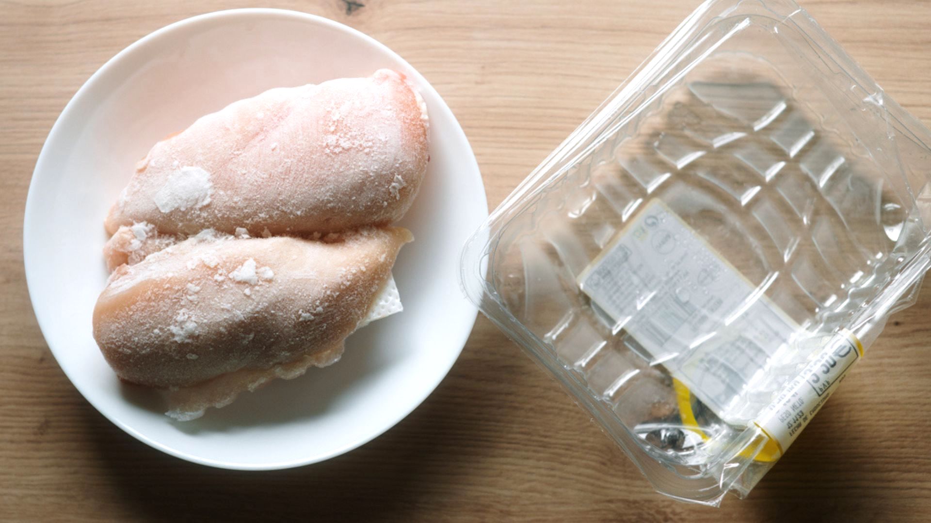 Two frozen chicken breasts on a white plate sit next to an empty clear plastic packaging on a wooden surface.