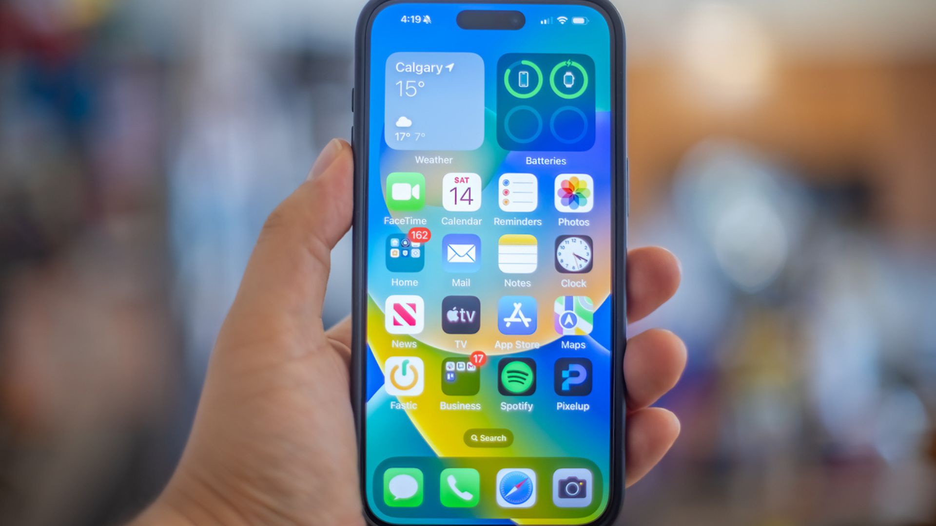 A hand holding an iPhone displaying the home screen with various app icons, including Weather, FaceTime, Calendar, and Maps. The screen shows Calgary weather at 15°C. The background is blurred.