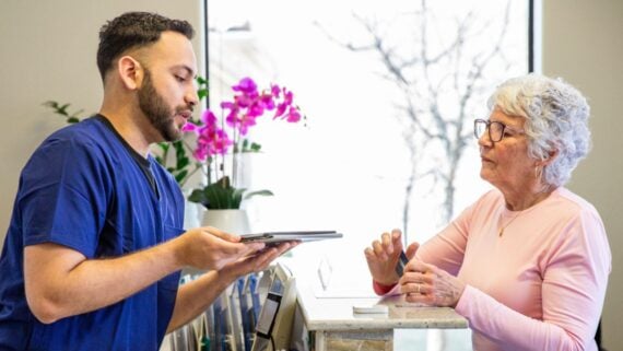 A healthcare professional in blue scrubs holds a tablet and speaks with an older woman at a reception desk. The woman wears glasses and a light pink top. A window and purple flowers are visible in the background.