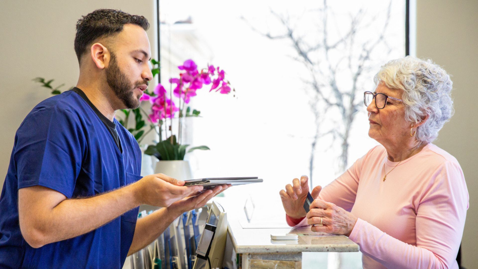 A healthcare professional in blue scrubs holds a tablet and speaks with an older woman at a reception desk. The woman wears glasses and a light pink top. A window and purple flowers are visible in the background.
