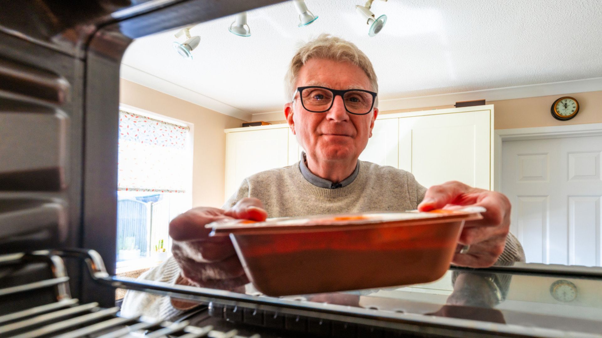 A man with glasses is placing a ready meal into an oven, viewed from inside the oven. The kitchen behind him is brightly lit, with white cabinets and a wall clock visible.