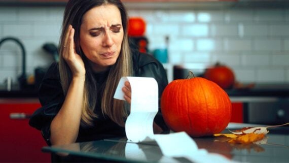A woman sits at a kitchen table, looking worried as she examines a long receipt. A pumpkin and autumn leaves are on the table, suggesting the fall season. The kitchen has white tile and red cabinets in the background.