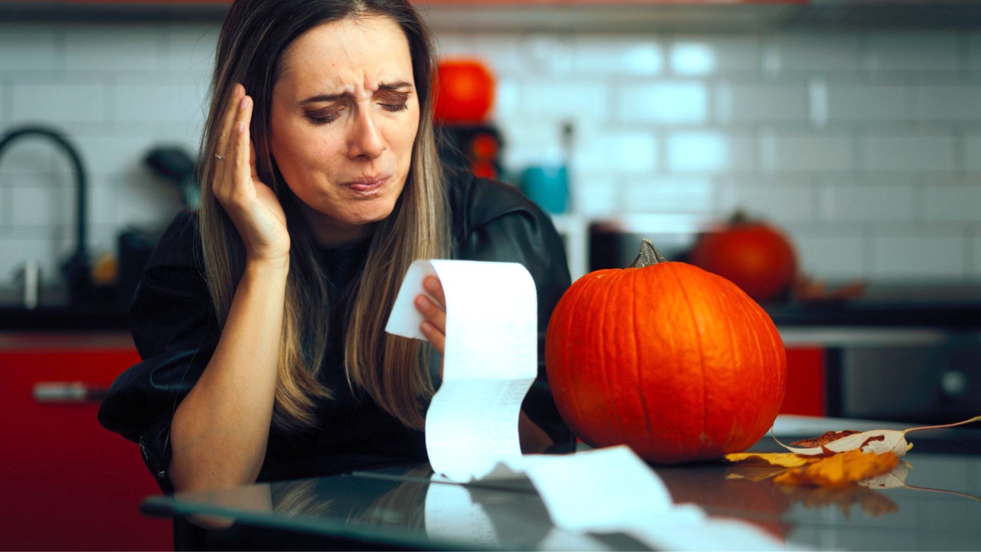 A woman sits at a kitchen table, looking worried as she examines a long receipt. A pumpkin and autumn leaves are on the table, suggesting the fall season. The kitchen has white tile and red cabinets in the background.