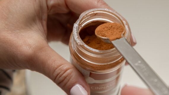 A hand holds an open jar of ground cinnamon while another hand scoops some out with a metal measuring spoon, carefully checking the label due to a recent cinnamon recall.