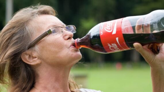 A woman with glasses drinks from a large bottle of Coca-Cola outdoors, with a blurred green park background.