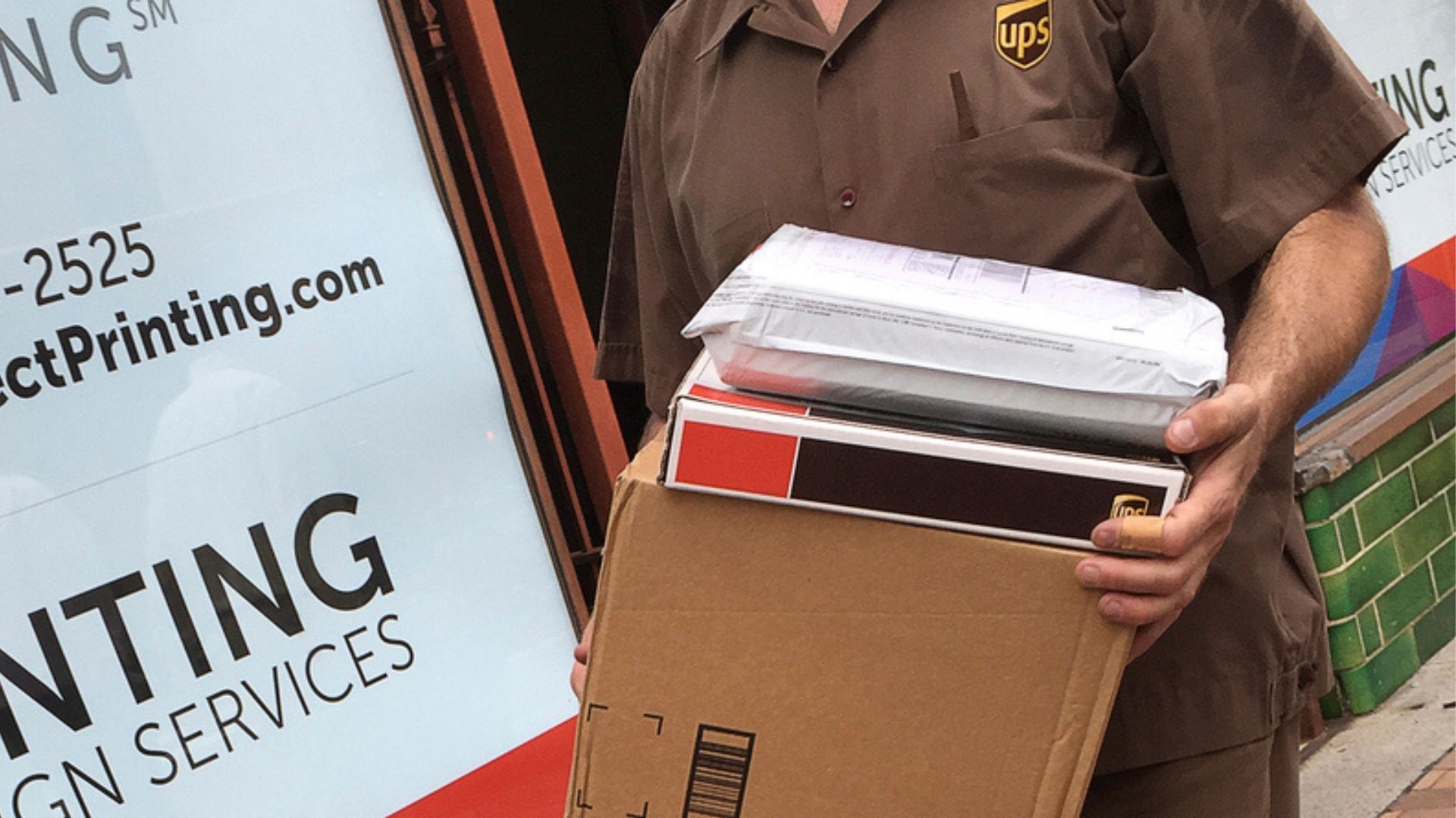 A UPS delivery worker in uniform holds a stack of packages, including a large cardboard box and two smaller parcels, standing outside a storefront with a partially visible sign.