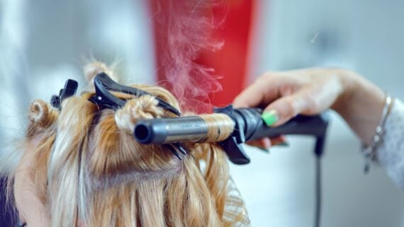 A close-up of a person curling blonde hair with a hot curling iron, with visible steam rising from the hair. The hand holding the iron has green nail polish.