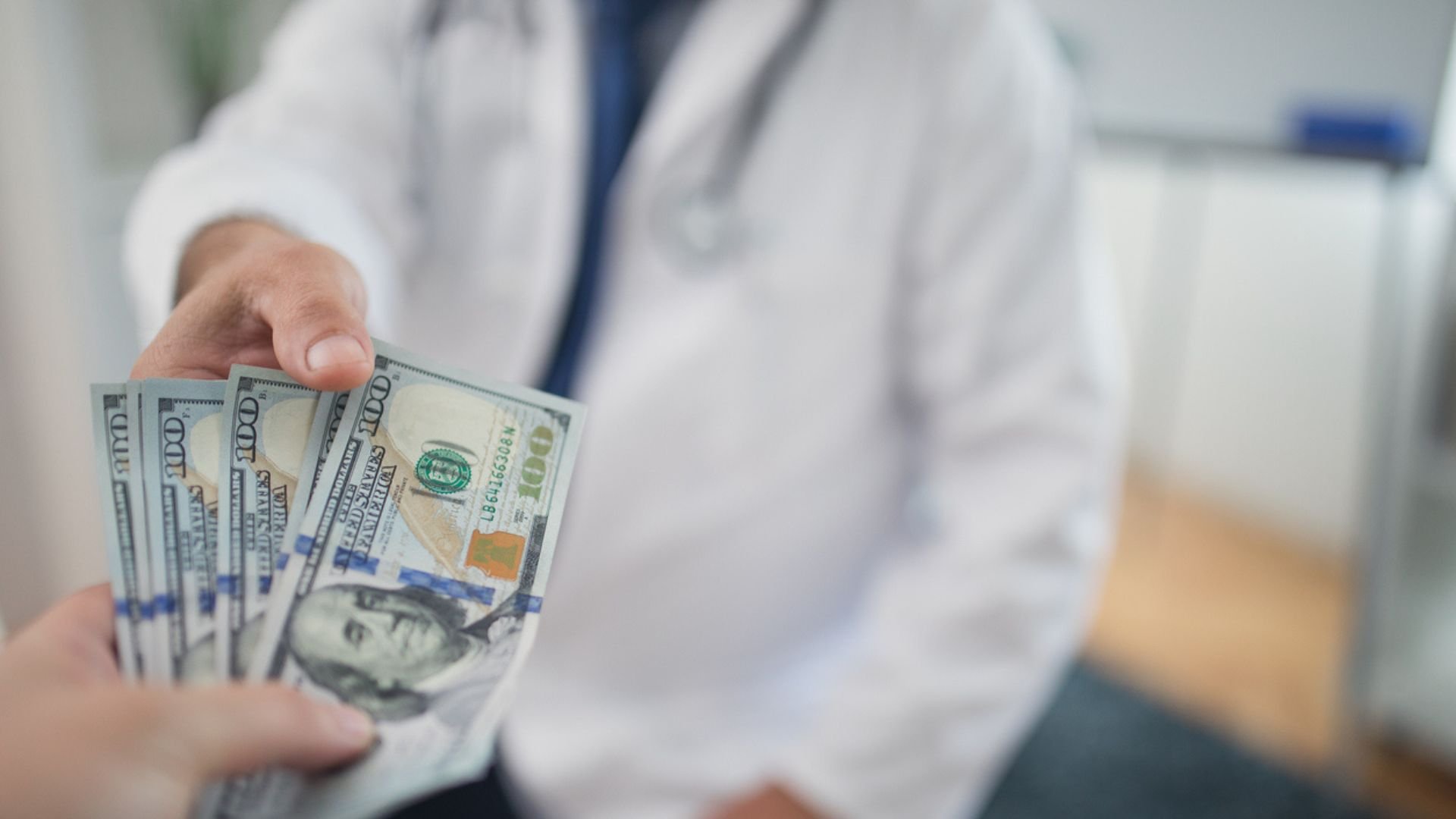 A person wearing a white lab coat, possibly a doctor, hands several $100 bills to another person. The focus is on the money being exchanged, with the background blurred.