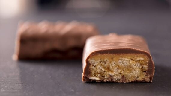 A close-up of a chocolate-covered candy bar cut in half, revealing a layered filling with nougat and caramel inside. Another whole piece is blurred in the background on a dark surface.