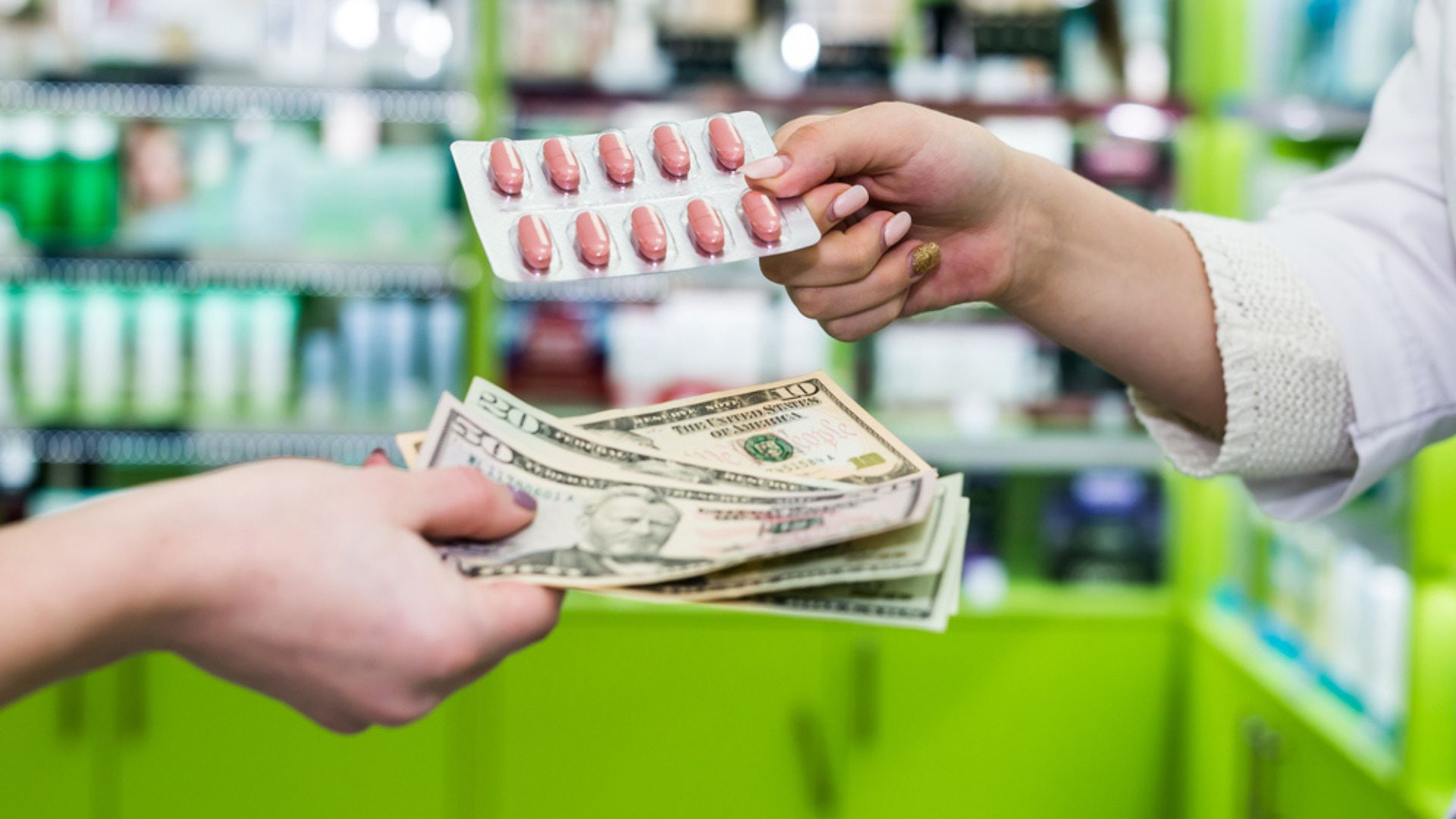 A person hands over paper currency while receiving a blister pack of pills from another person across a pharmacy counter with shelves filled with products in the background.