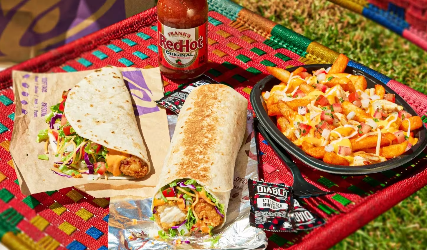 Two loaded Taco Bell burritos, seasoned fries topped with sauce and diced vegetables, hot sauce packets, and a bottle of Frank's RedHot sauce on a colorful woven table outdoors.