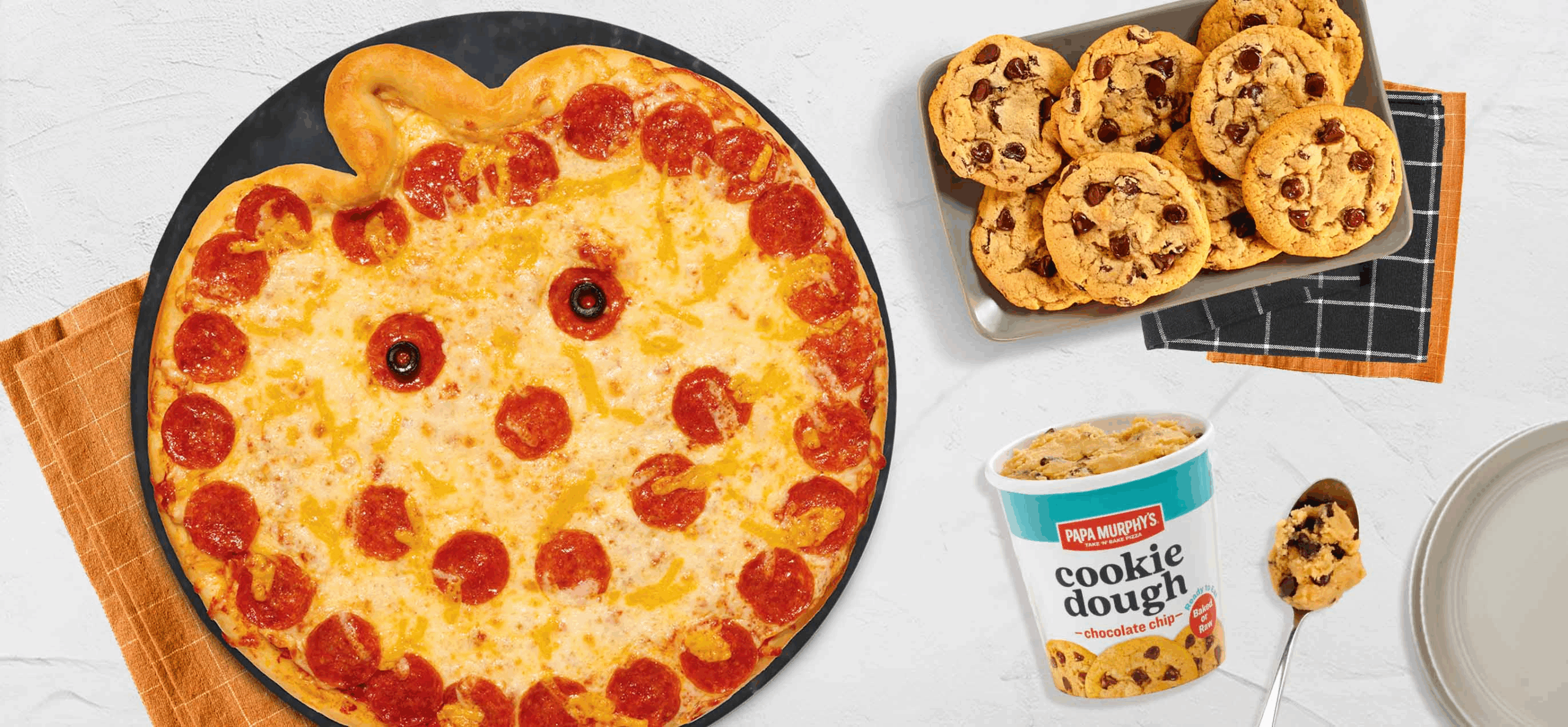 A pumpkin-shaped pepperoni pizza with a smiling face is on a black platter, next to a tray of chocolate chip cookies, a tub of Papa Murphy's cookie dough, and a spoon with cookie dough on it.
