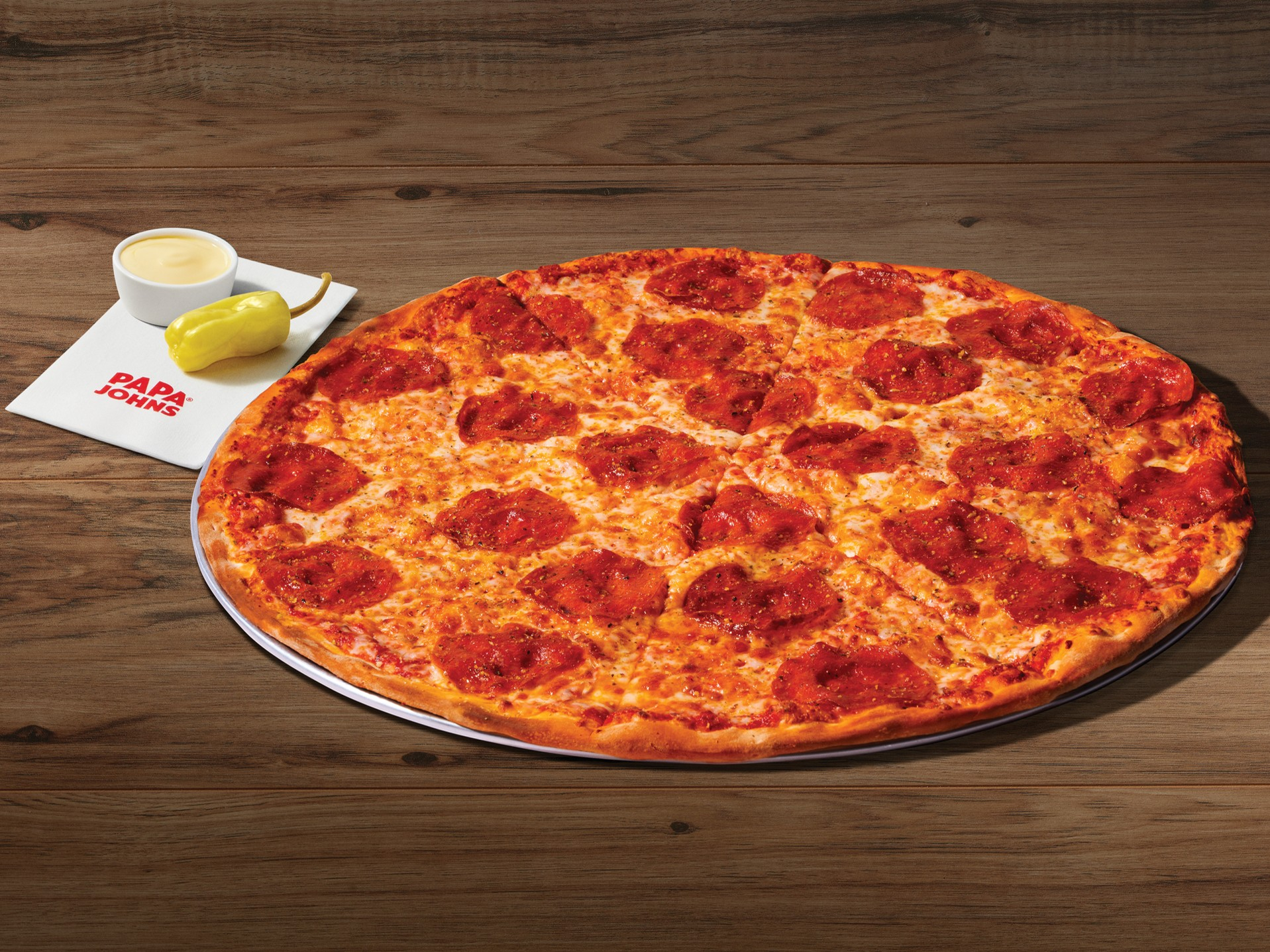 A large pepperoni pizza from the new fast-food menu sits on a round tray atop a wooden table, accompanied by a pepperoncini pepper, garlic sauce cup, napkin, and a pizza box in the background. grand papa 