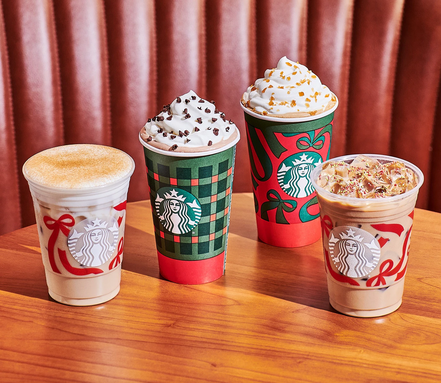 Four Starbucks holiday-themed drinks from the new fast-food menu are displayed on a wooden table; two are iced, and two are topped with whipped cream and festive sprinkles. The cups feature seasonal red and green designs, with a brown cushioned booth in the background.