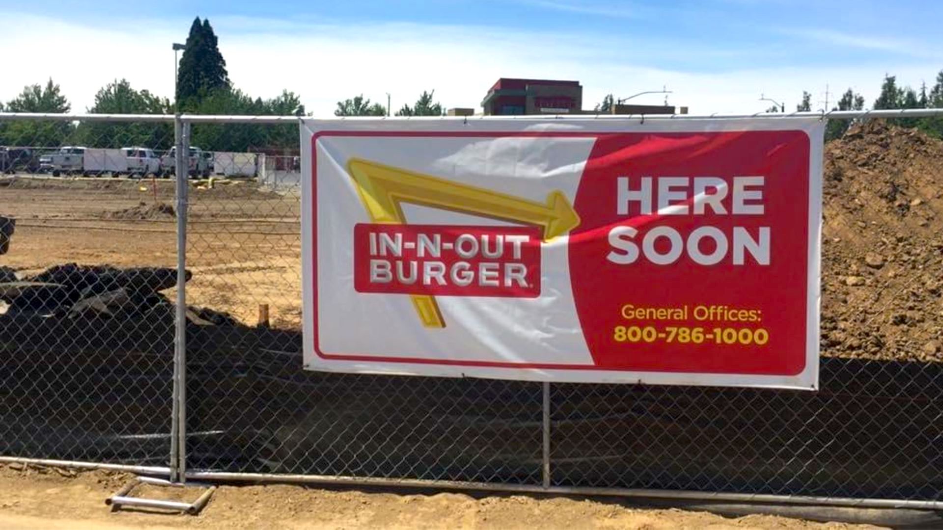 A large banner on a construction fence reads “IN-N-OUT BURGER HERE SOON” with a phone number and logo, in front of a dirt lot and building site under a blue sky.