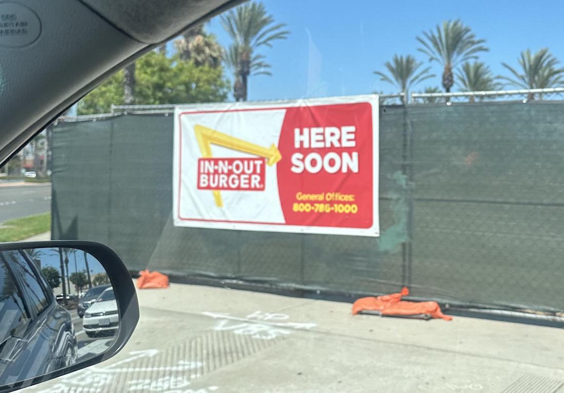 A construction site is visible behind a green fence with a large sign that reads "IN-N-OUT BURGER. HERE SOON. General Offices: 800-786-1000," seen from a car window. Palm trees line the background.
