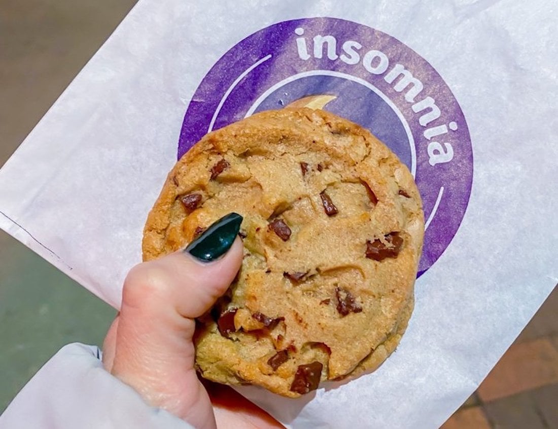 A hand with dark-painted nails holds a chocolate chip cookie in front of an Insomnia Cookies branded paper bag.