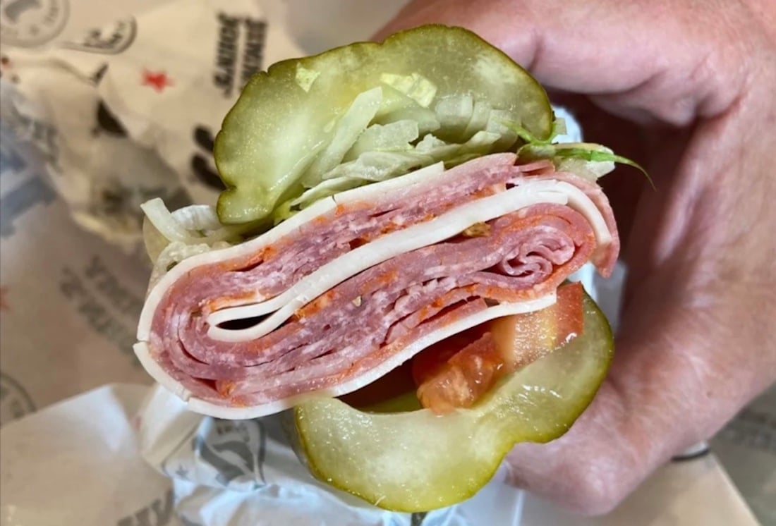 A hand holding a sandwich made with pickle halves instead of bread, filled with layers of deli meats, shredded lettuce, tomato, and cheese.