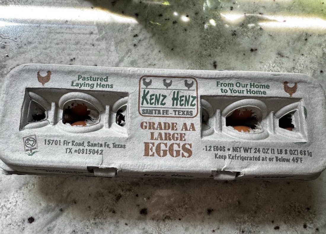 A carton of Kenz Henz Grade AA large eggs sits on a countertop. The carton is labeled as containing 12 eggs from pastured laying hens, produced in Santa Fe, Texas. Some eggs are visible through the openings.
