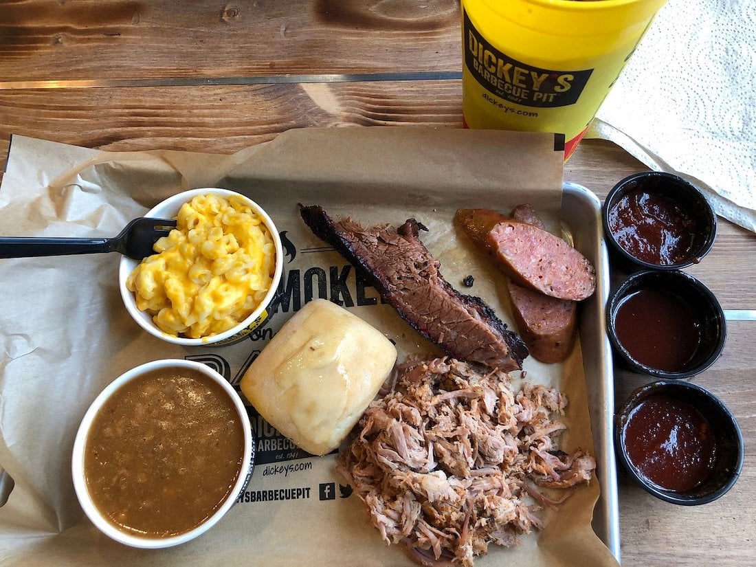 A barbecue meal on a tray with macaroni and cheese, baked beans, a bread roll, sliced brisket, sausage, pulled pork, three cups of barbecue sauce, and a large yellow Dickey’s Barbecue Pit drink cup.