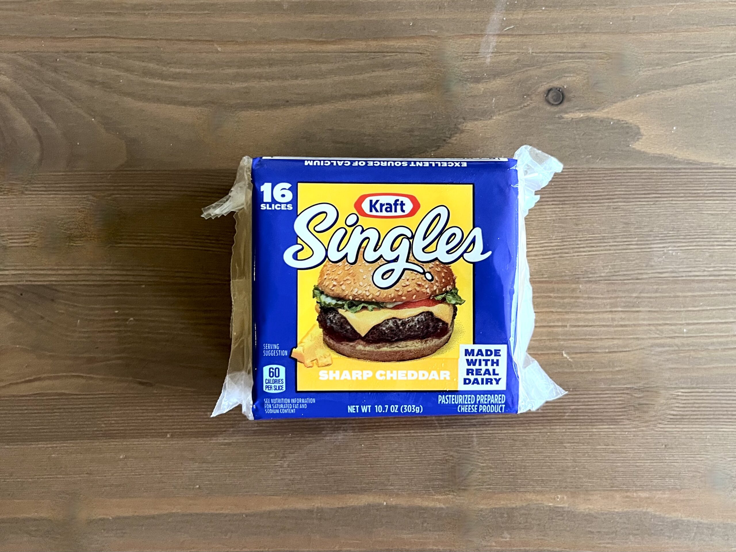 A package of Kraft Singles Sharp Cheddar cheese slices sits on a wooden surface. The blue package features an image of a cheeseburger and states it contains 16 slices and is made with real milk.