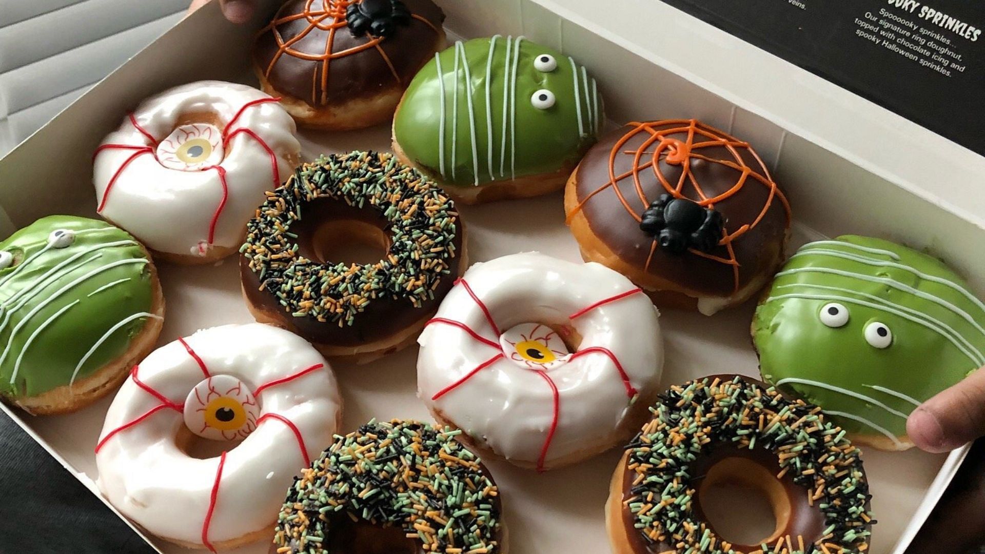 A box of Halloween-themed donuts decorated with green icing, candy eyes, orange and black spider webs, and green and orange sprinkles. Some donuts have monster faces or eyeball designs on white icing.