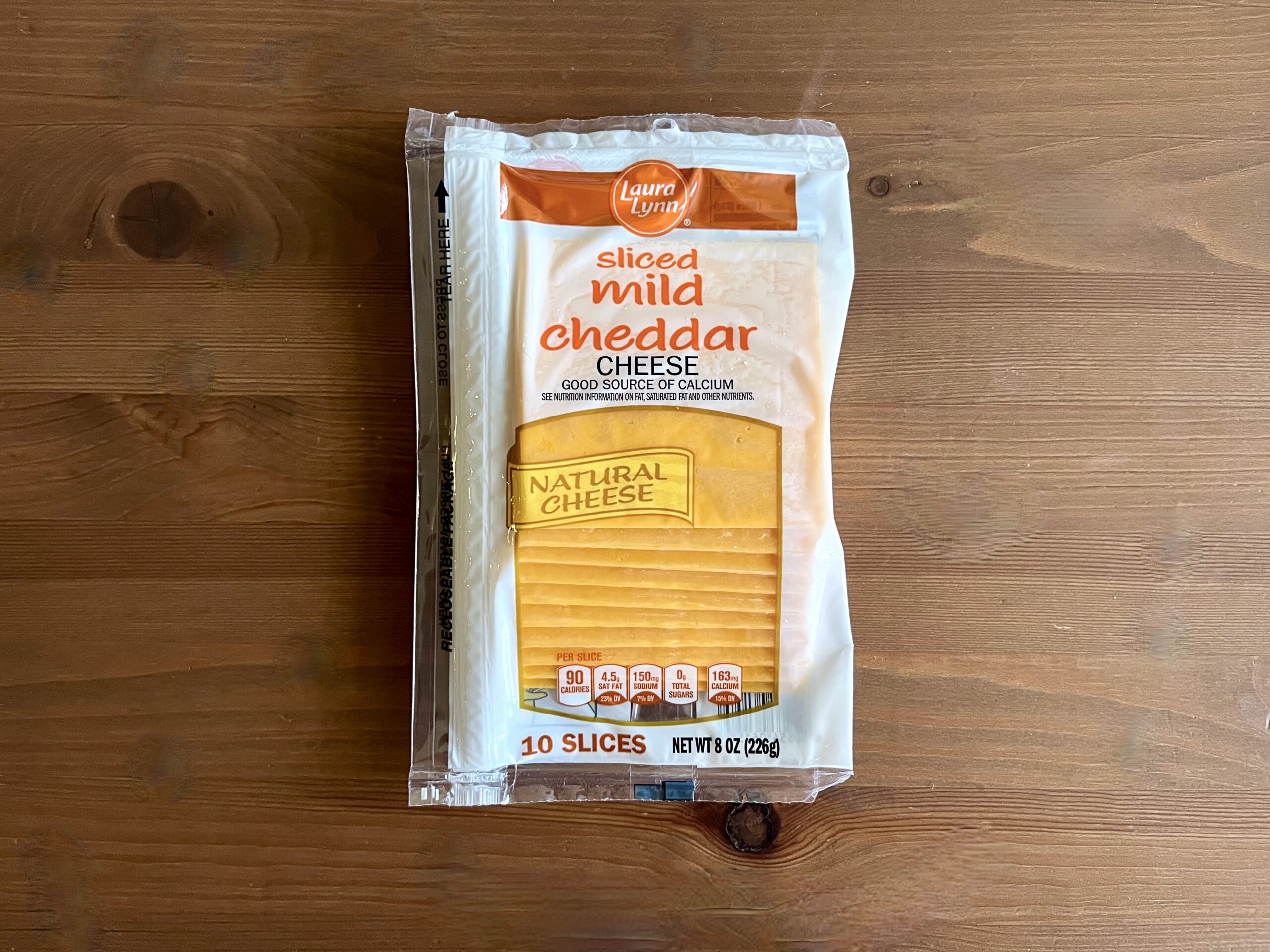 A package of Laura Lynn sliced mild cheddar cheese sits on a wooden surface. The clear packaging reveals 10 slices of yellow cheese, and the label highlights it as a good source of calcium.