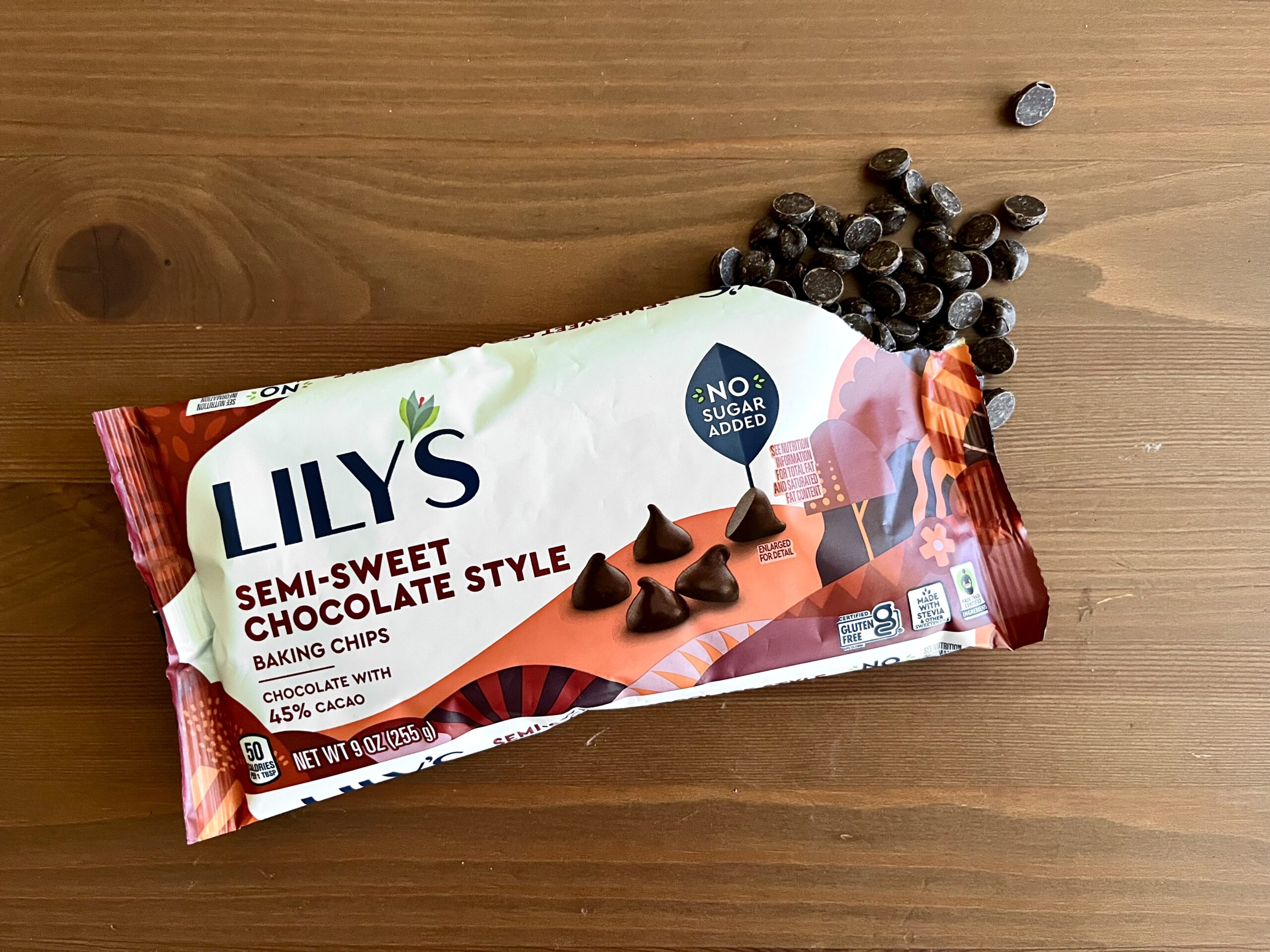 A package of Lily’s semi-sweet chocolate style baking chips rests on a wooden surface, with some chocolate chips scattered nearby. The packaging highlights “No Sugar Added” and contains images of chocolate chips.