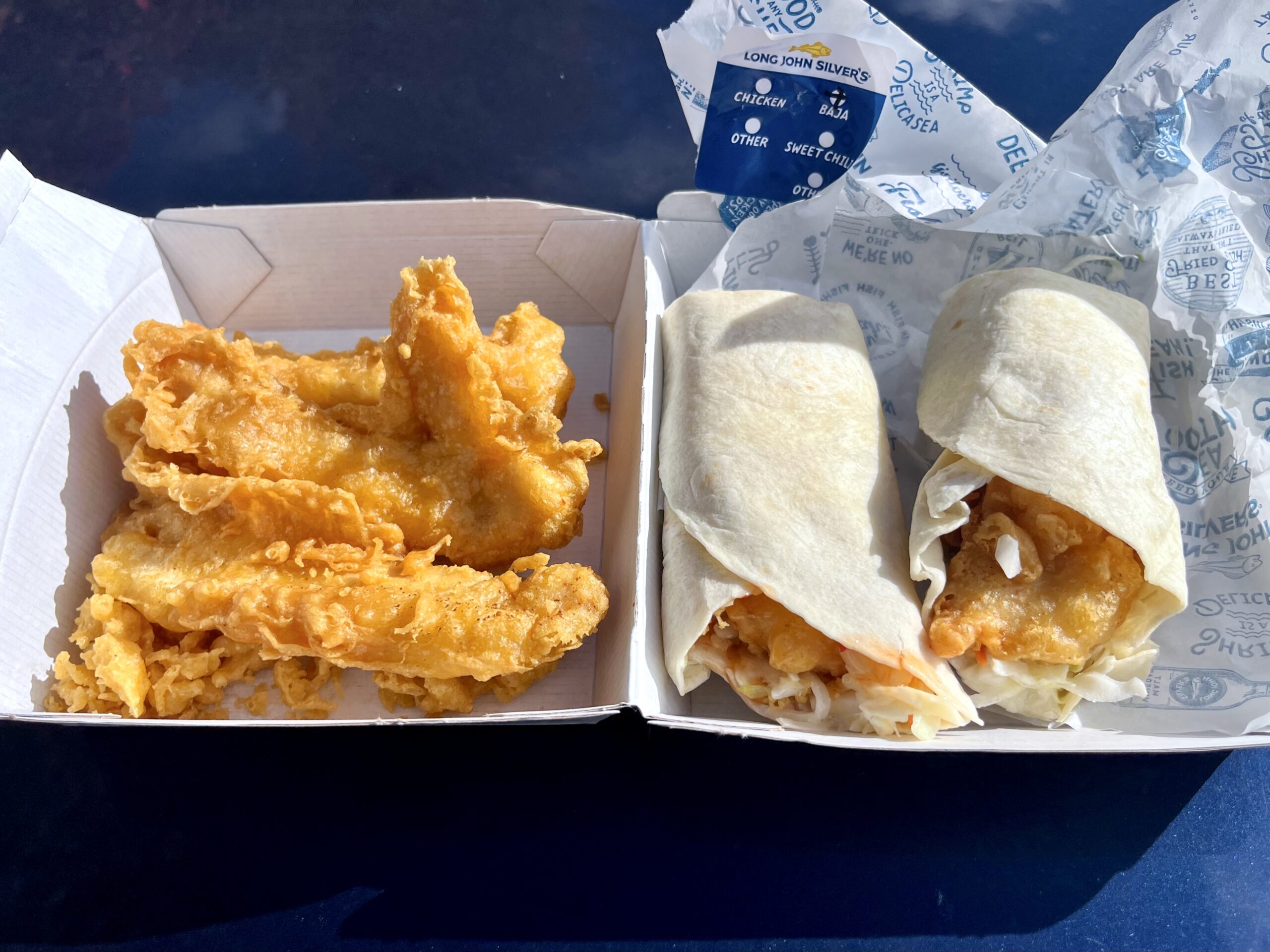 Two pieces of fried fish in a white box next to two fish tacos wrapped in tortillas with shredded lettuce, all served on a blue surface.