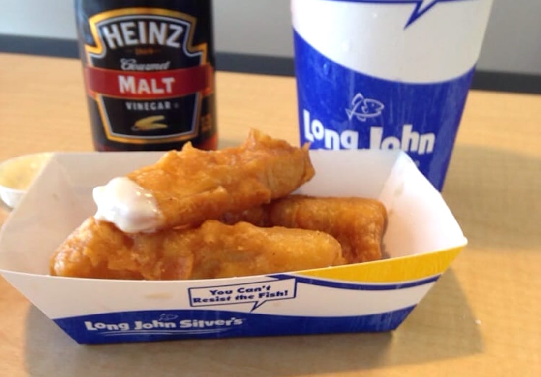 A tray of battered fish sticks with tartar sauce, a bottle of Heinz malt vinegar, and a blue Long John Silver's drink cup are placed on a light brown table.