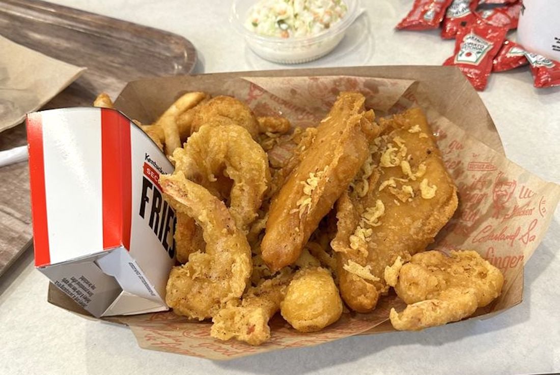 A basket of fried food including shrimp, fish fillets, and fries, served in a paper tray with a box labeled "FRIES." In the background are a small cup of coleslaw and several ketchup packets.