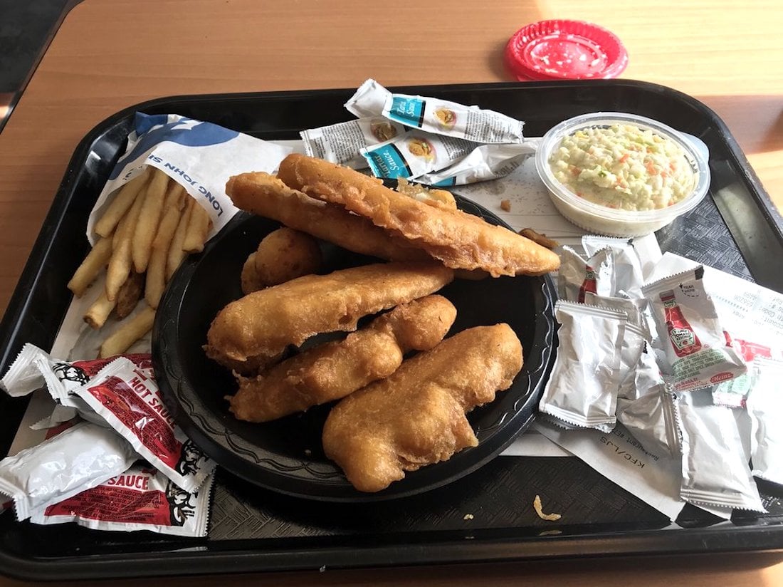A black tray with fried fish fillets, hush puppies, French fries, coleslaw in a plastic cup, and assorted condiment packets on a table.
