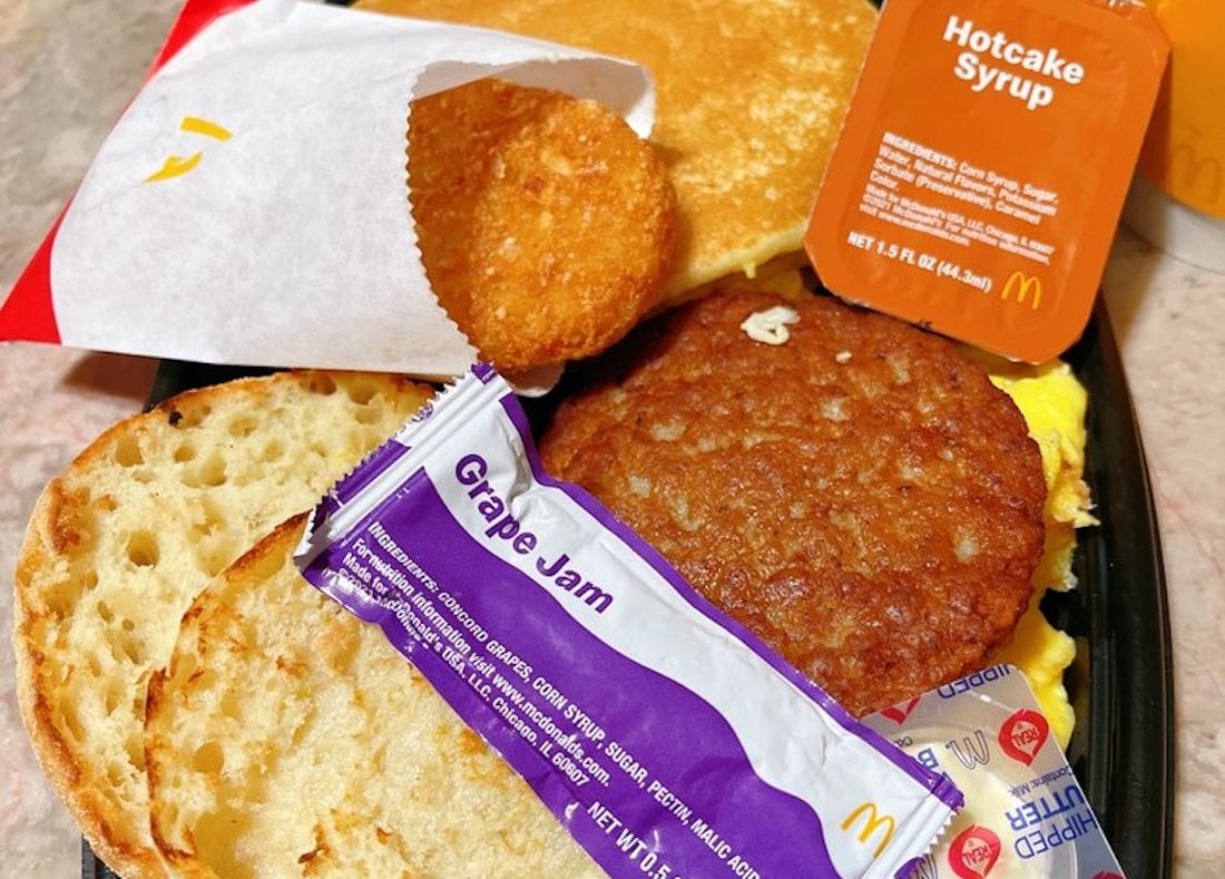 A McDonald's breakfast with an English muffin, sausage patty, scrambled eggs, hash brown, hotcake, grape jam packet, and a cup of hotcake syrup on a black tray.