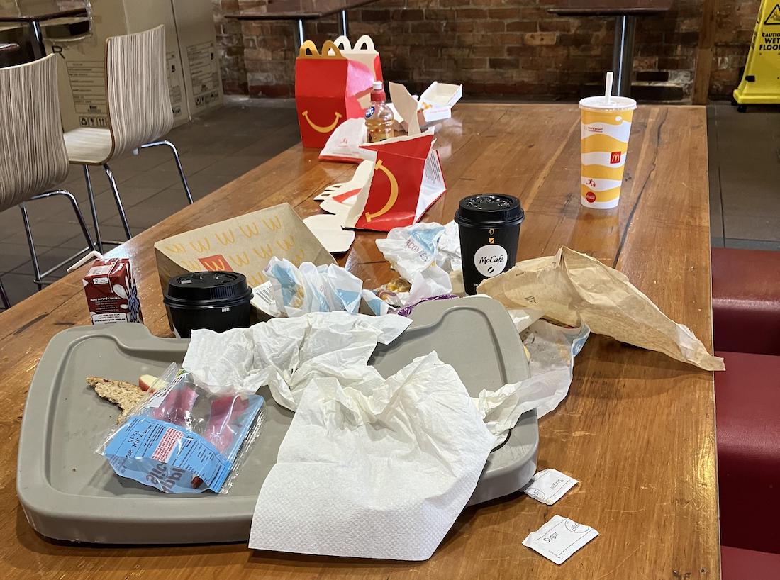 A messy restaurant table covered with empty fast food packaging, used napkins, trays, drink cups, and food wrappers. In the background are empty chairs, tables, and a brick wall.