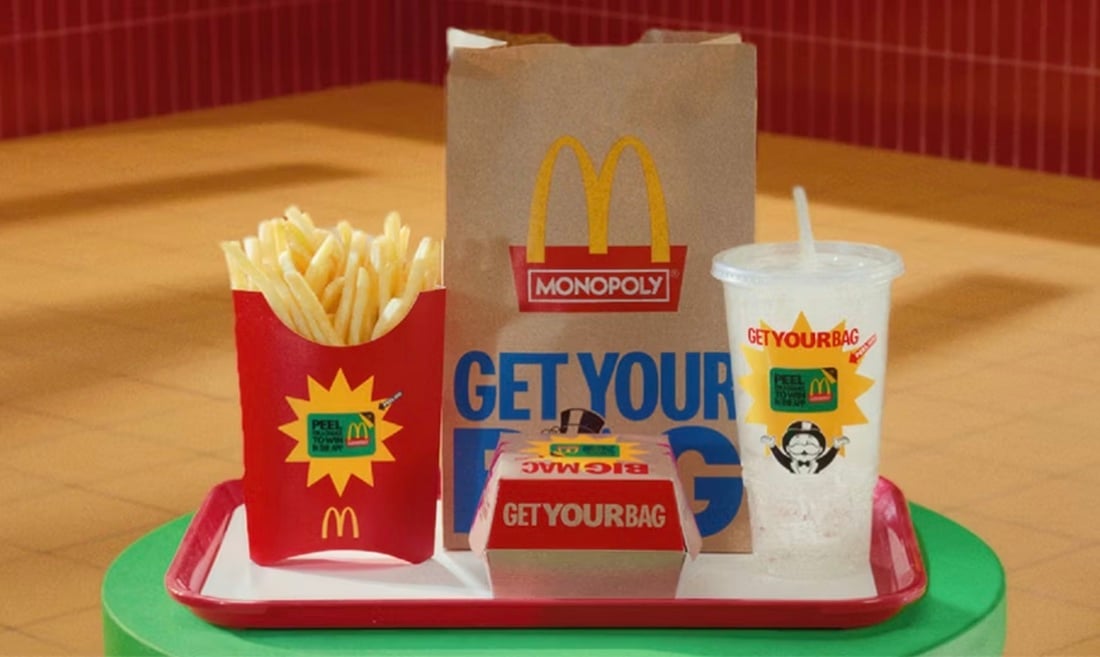A McDonald’s meal on a red tray includes fries, a Big Mac in a box, a soft drink, and a brown paper bag. All packaging displays Monopoly-themed promotional stickers. The background is a tiled wall and floor.